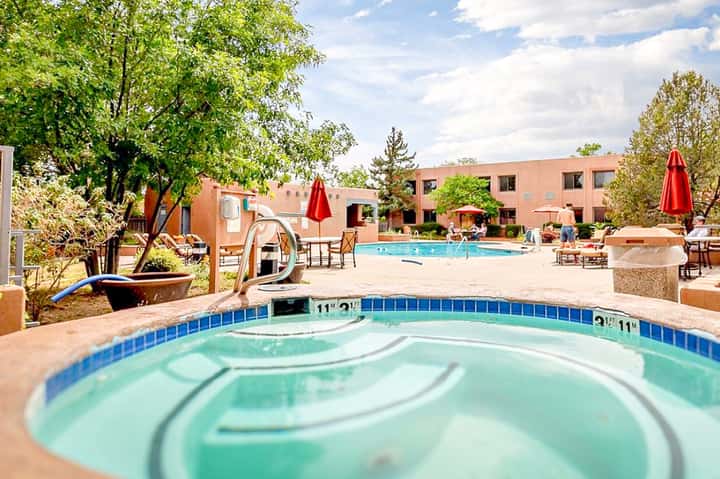 Resort pool and hot tub surrounded by lounge chairs, red umbrellas, trees, and terracotta buildings