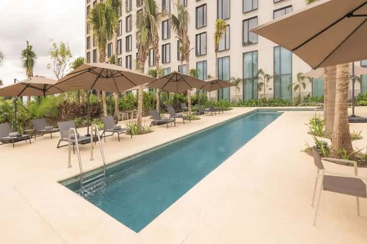 Modern hotel pool deck with lap pool, tan umbrellas, lounge chairs, tropical plants, and tall residential building in background