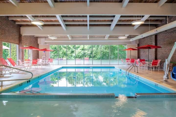 Indoor pool with blue water, spa area, red umbrellas, outdoor seating, and large windows