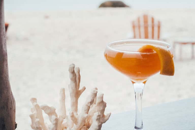 Amber cocktail with orange slice garnish on beach with coral backdrop