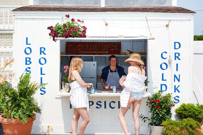 Beachside cocktail bar window with blue text signage and two women in white outfits ordering drinks