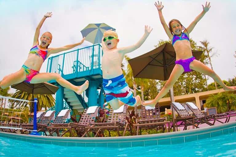 Three children jumping into a swimming pool with umbrellas and loungers in background
