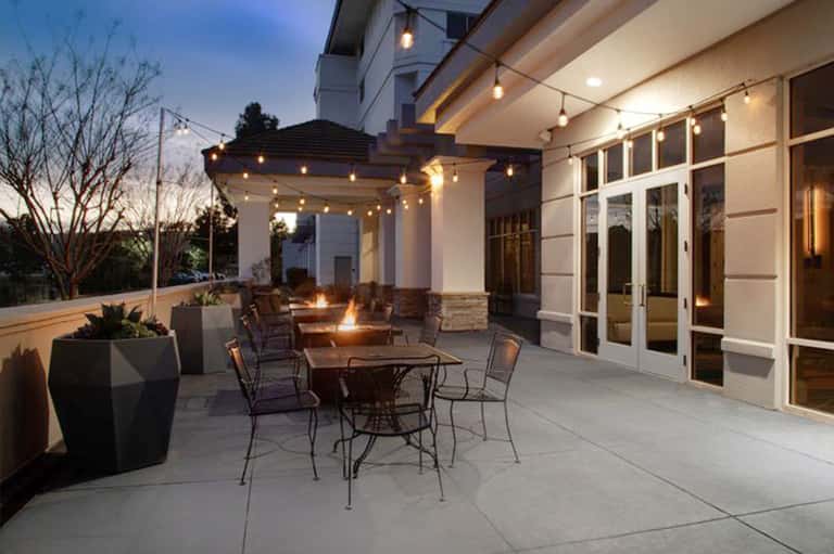 Modern patio with fire pit, wooden pergola, string lights, and contemporary furnishings at dusk