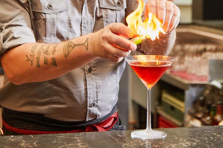 Bartender igniting a flaming cocktail with lime garnish at bar counter