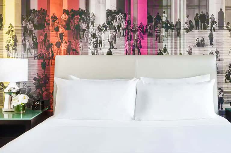 Modern hotel bedroom with white bedding and colorful digital art mural of crowded public space