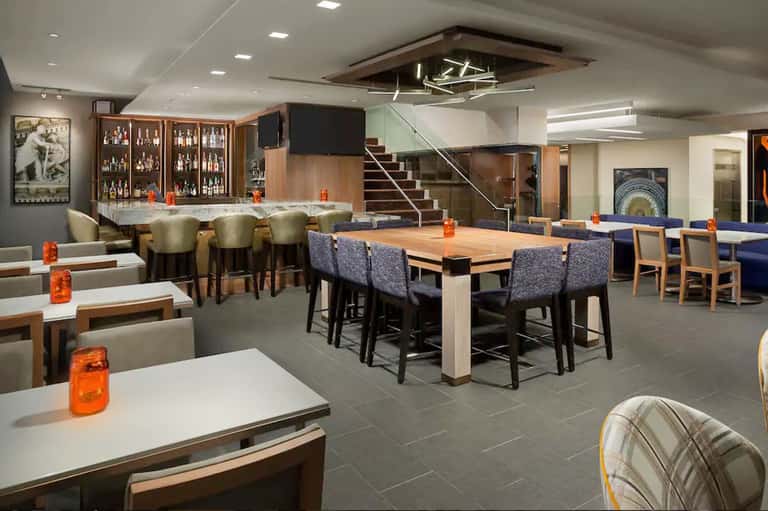 Modern hotel lounge with bar, dining tables, and staircase to upper level