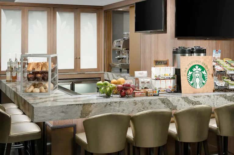 Hotel breakfast area with marble countertop, Starbucks signage, pastries, fresh fruit, and wood-paneled walls