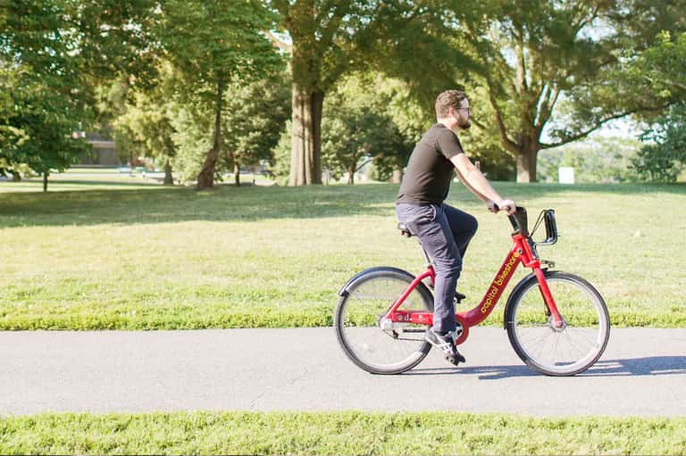Man riding red bike on paved path through tree-lined park on sunny day