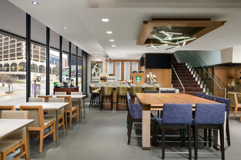 Modern café interior with bar seating, wooden tables, large windows, and contemporary staircase