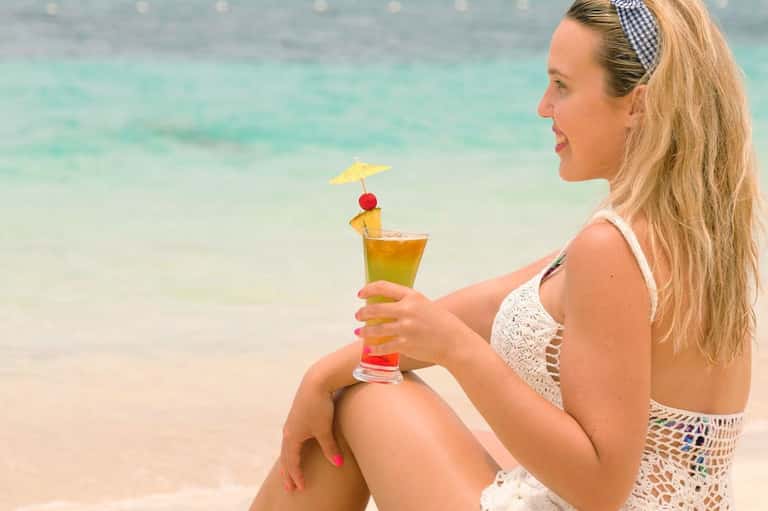 Woman in white swimsuit enjoying tropical cocktail on beach with turquoise ocean water