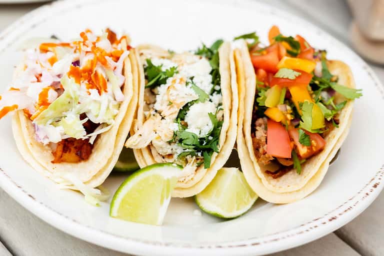 Three fish tacos on white plate with lime wedges, cilantro, and colorful peppers