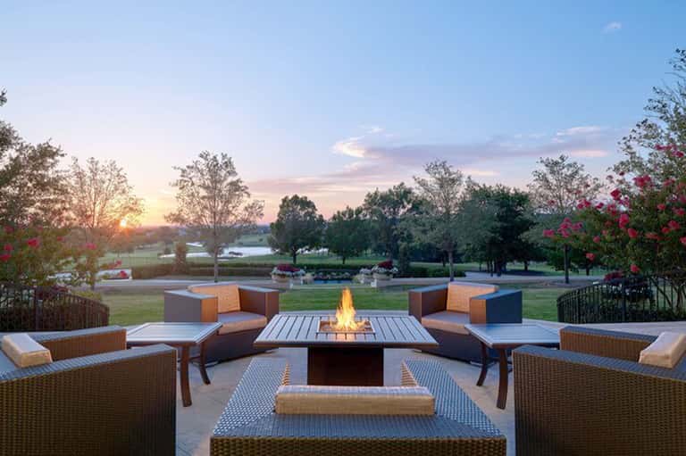 Fire pit seating area with woven chairs and pink flowers overlooking a sunset landscape