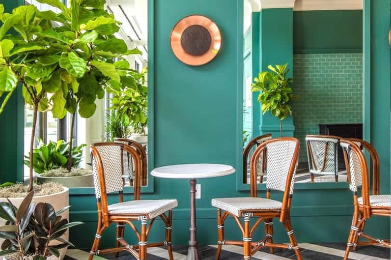 Turquoise outdoor café with rattan chairs, small round table, potted plants, and coral-colored speaker on wall
