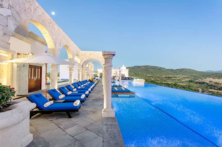 Infinity pool with blue loungers under white arches overlooking desert landscape