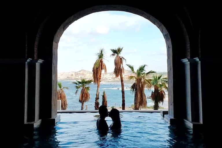 Couple in infinity pool framed by archway, palm trees, and turquoise ocean vista beyond