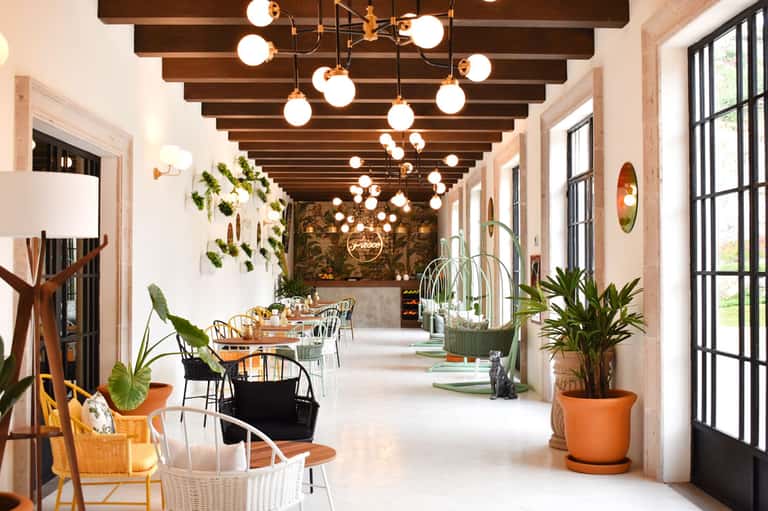 Modern restaurant corridor with exposed wood beams, globe pendant lights, potted plants, and dining tables along large windows