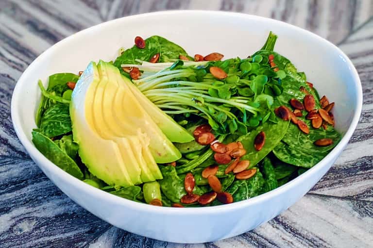Fresh green salad bowl with avocado slices, mixed greens, and red pepper flakes on striped cloth