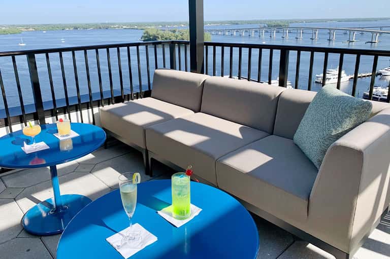 Waterfront balcony with gray upholstered sofa, blue side tables, and bridge views over water