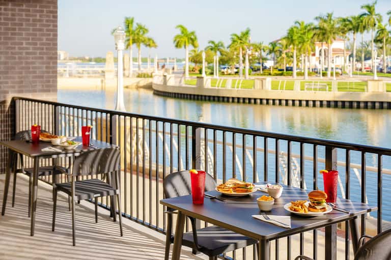 Waterfront dining terrace with tables overlooking a scenic river and palm trees in tropical setting