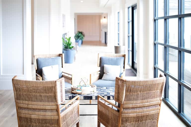 Modern lounge area with woven chairs, blue accents, and bright windows overlooking hallway