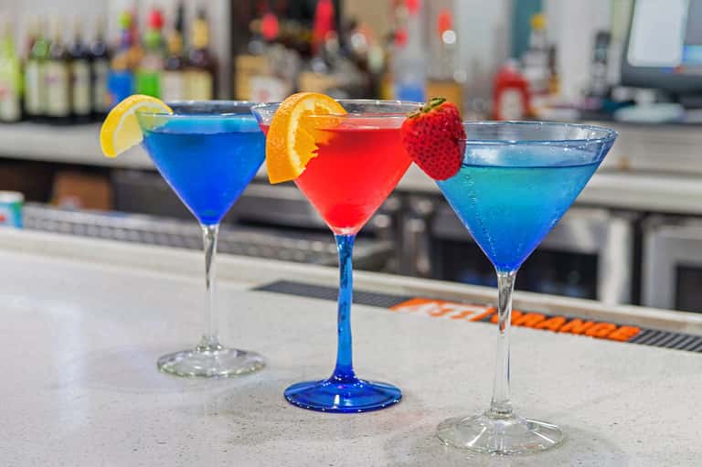 Three colorful cocktails on a bar counter with fruit garnishes and blue, red, and tropical drinks