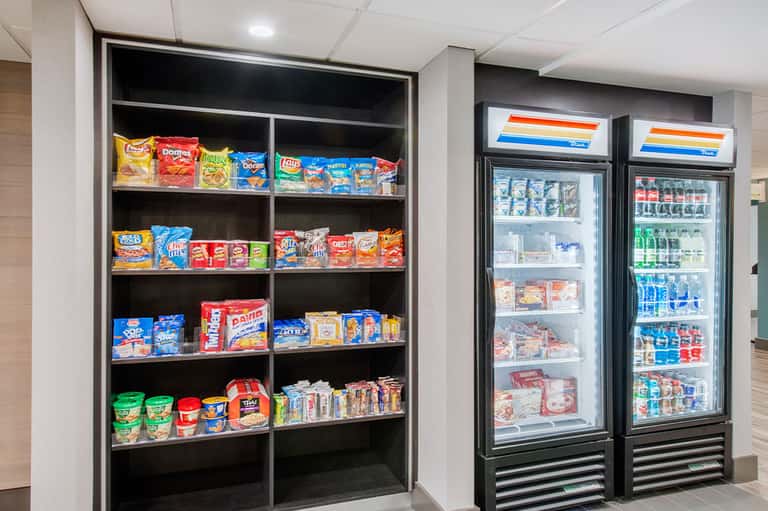 Hotel snack bar with open shelving displaying chips and packaged goods, two refrigerated beverage coolers with drinks