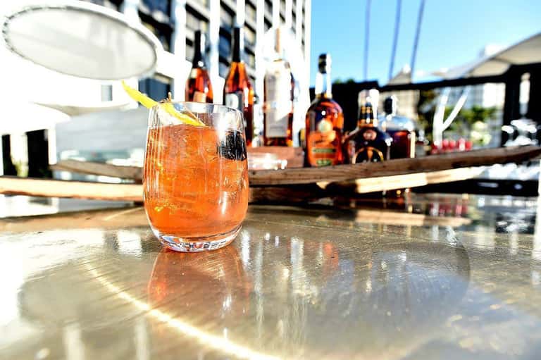 Cocktail with orange garnish on outdoor bar counter with bottles and staff in background