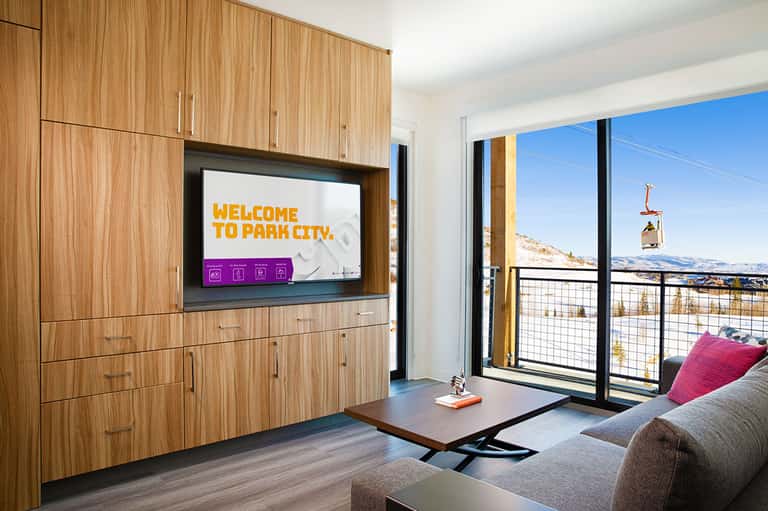 Modern ski resort condo interior with wood cabinetry, TV, and balcony overlooking snowy mountains and ski lift