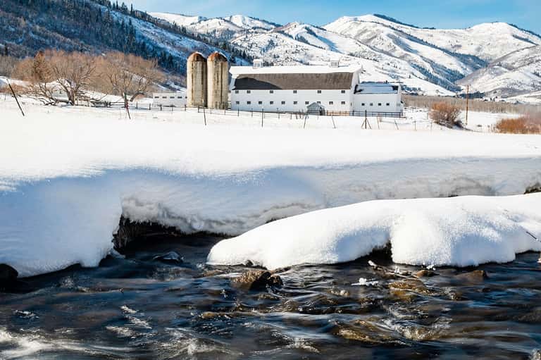 Snow-covered farmland with a white barn and silos against snow-capped mountains