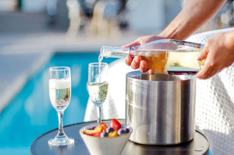 Hands pouring champagne by a poolside with glasses and fruit bowl