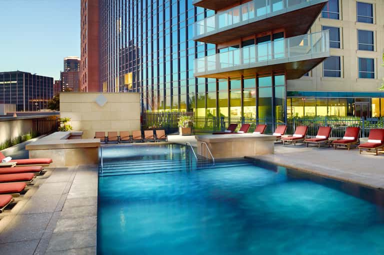 Modern rooftop pool with turquoise water, red lounge chairs, and city skyline with tall buildings