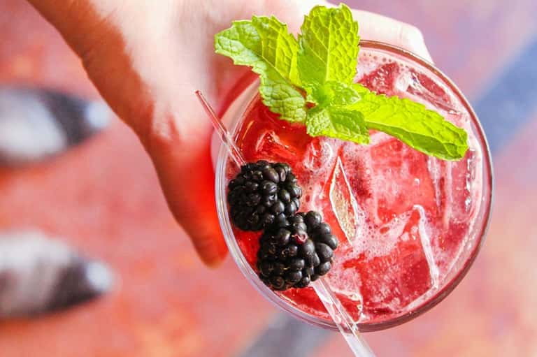 Red cocktail with ice, fresh blackberries, mint leaves, and straw in clear glass