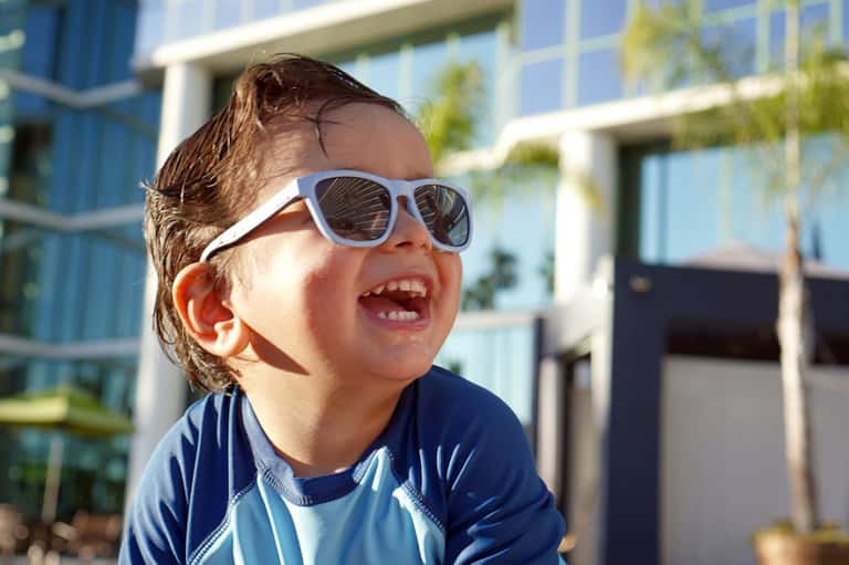 Young child wearing white sunglasses and blue rash guard smiling in sunny resort courtyard