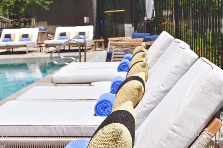 Poolside lounge chairs with white cushions, blue rolled towels, and straw hats arranged in a row by the pool