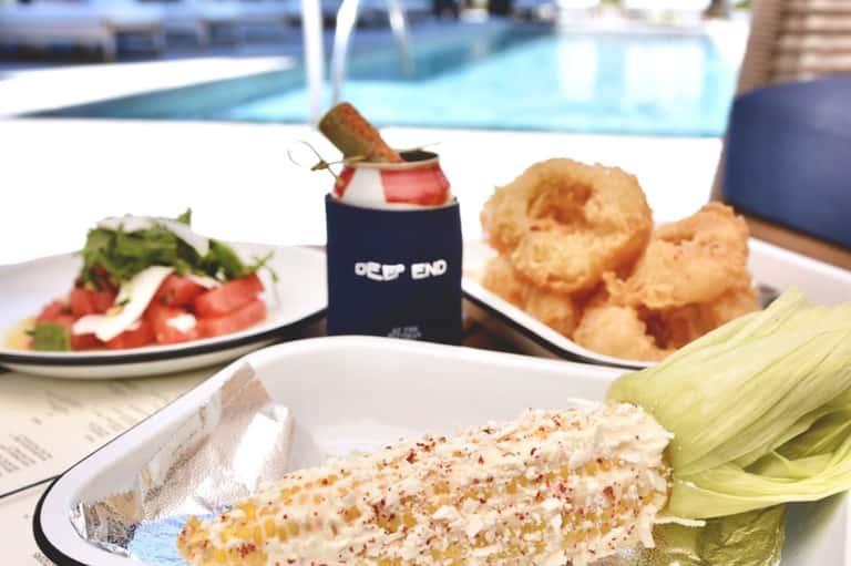 Poolside dining spread with grilled corn, salad, fried appetizers, cocktail, and scenic pool view in background