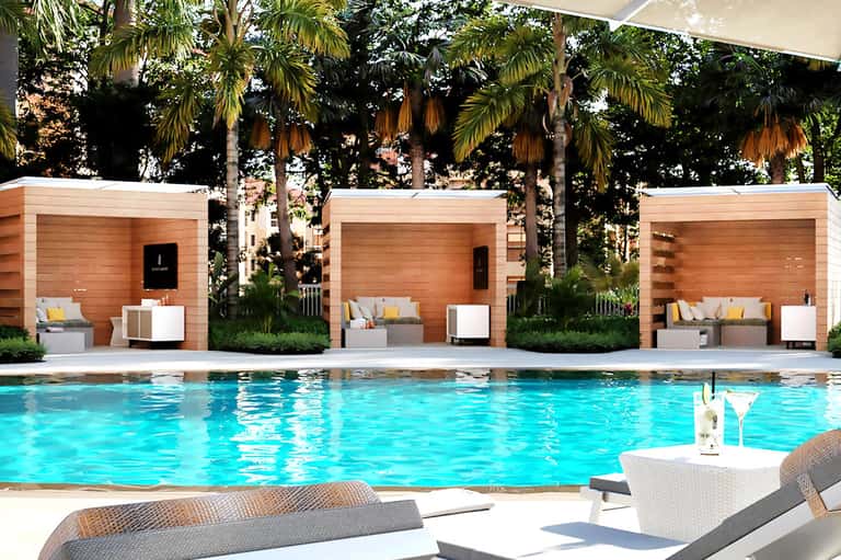 Luxury resort pool with turquoise water, wooden cabanas, palm trees, and loungers