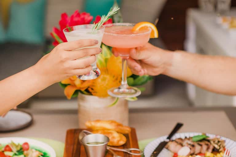 Two cocktails clinking together poolside with fresh flowers and appetizers nearby