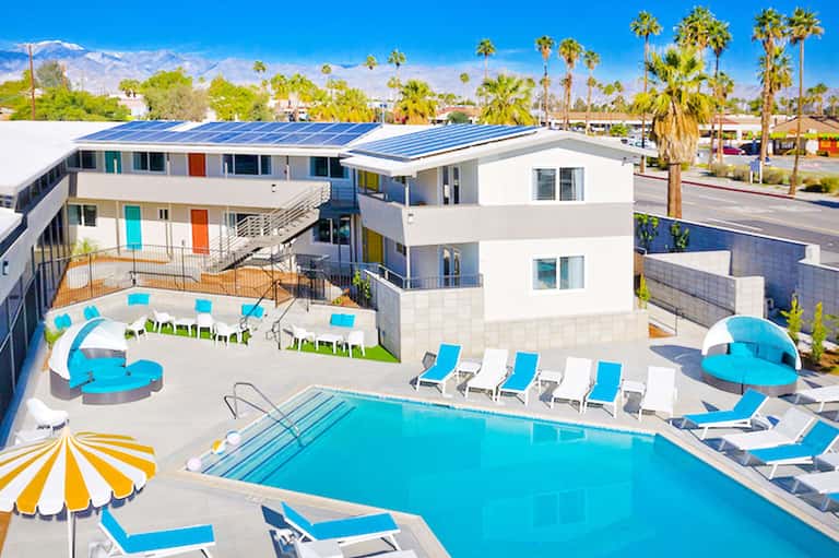 Modern resort courtyard with swimming pool, blue loungers, shade umbrellas, and multi-story white building