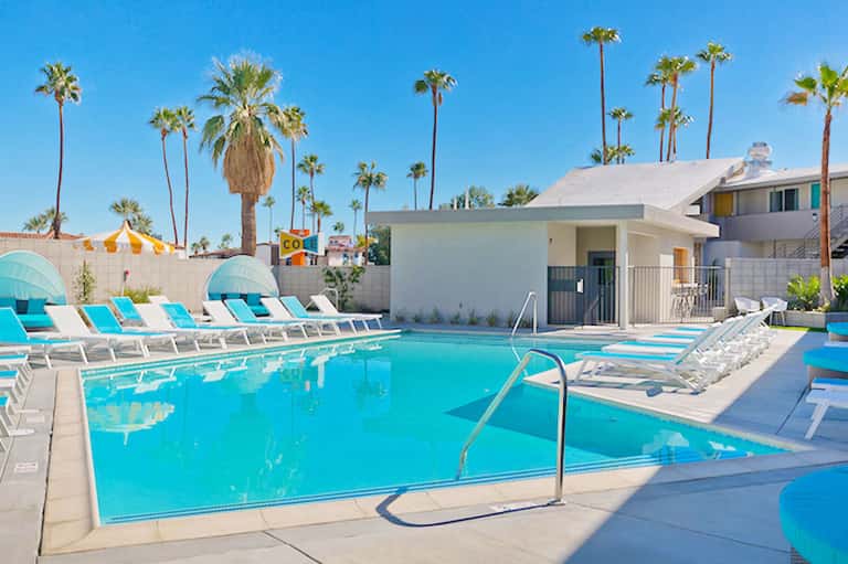 Bright turquoise swimming pool with lounge chairs, umbrellas, and palm trees under blue sky