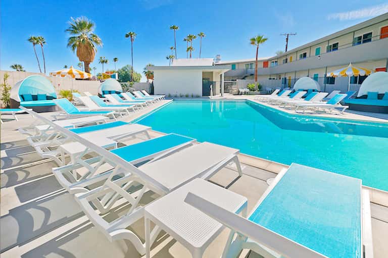 Turquoise swimming pool with white lounge chairs and blue cabanas surrounded by palm trees