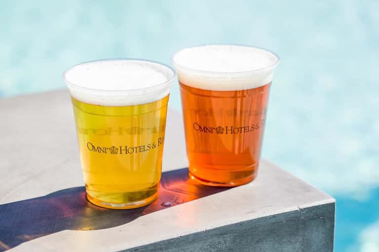 Two Omni Hotels branded beers with white foam heads on a poolside ledge against blue water