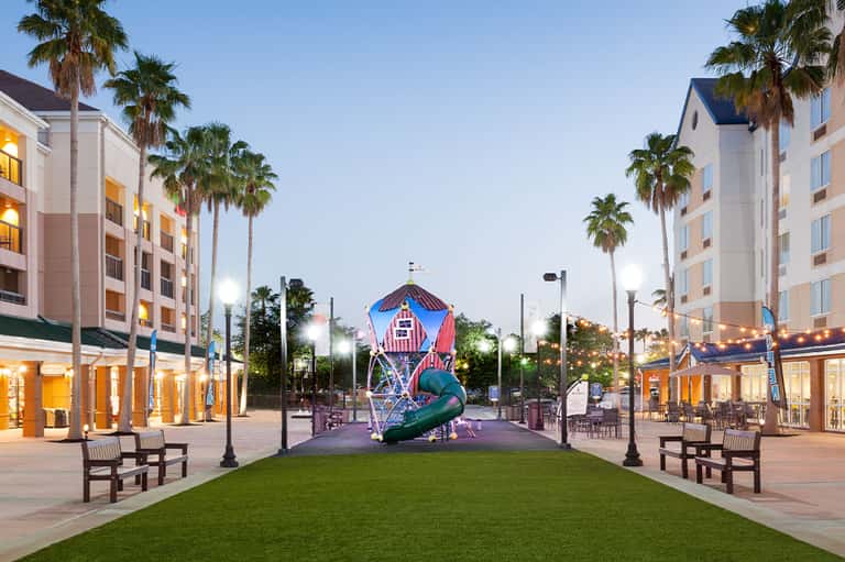 Hotel courtyard at dusk with illuminated buildings, palm trees, and colorful playground sculpture