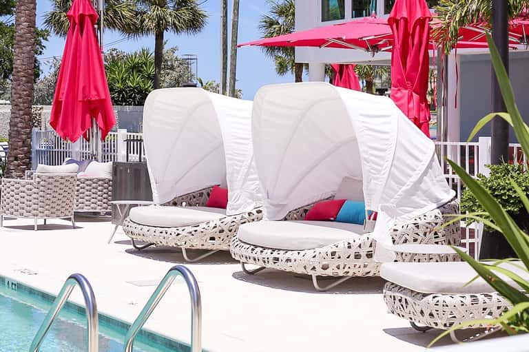Luxury poolside cabanas with white canopies and cushions, pink umbrellas, palm trees, and tropical landscaping