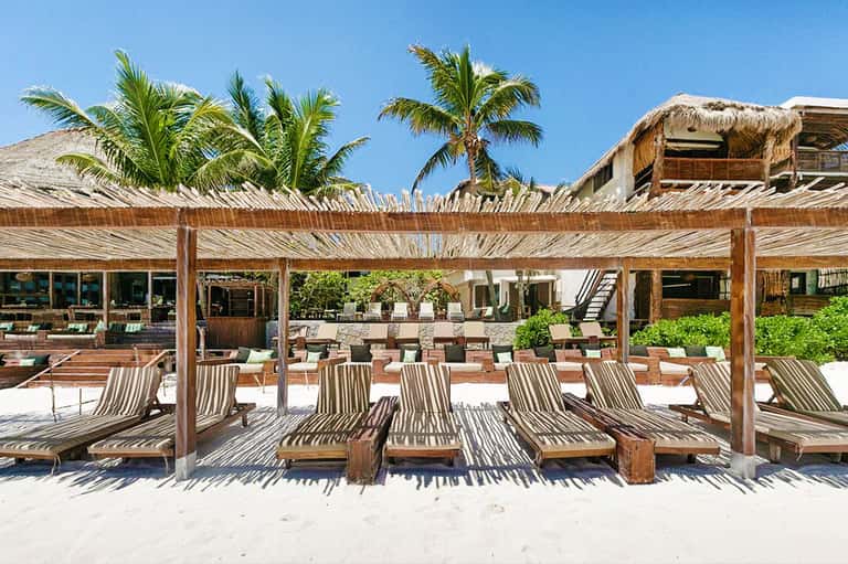Beach club with striped lounge chairs under wooden pergolas, palm trees, and thatched-roof structures