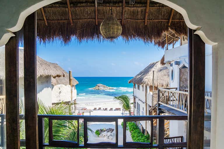 Beachfront bungalow view framed by thatched roof, overlooking turquoise ocean and white sand beach