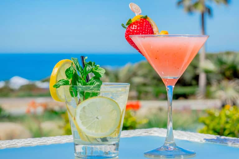 Tropical cocktails with fresh fruit garnishes on a blue table overlooking the ocean and palm trees