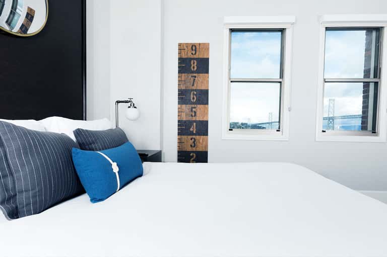 Modern hotel bedroom with white bedding, blue pillows, and bay bridge view through windows