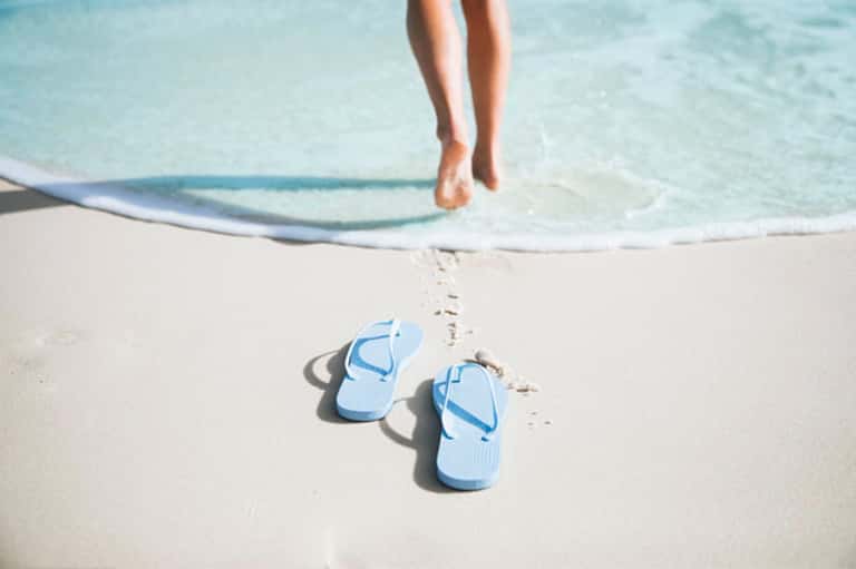 Pair of blue flip-flops on white sand beach with ocean waves and surfboard in background
