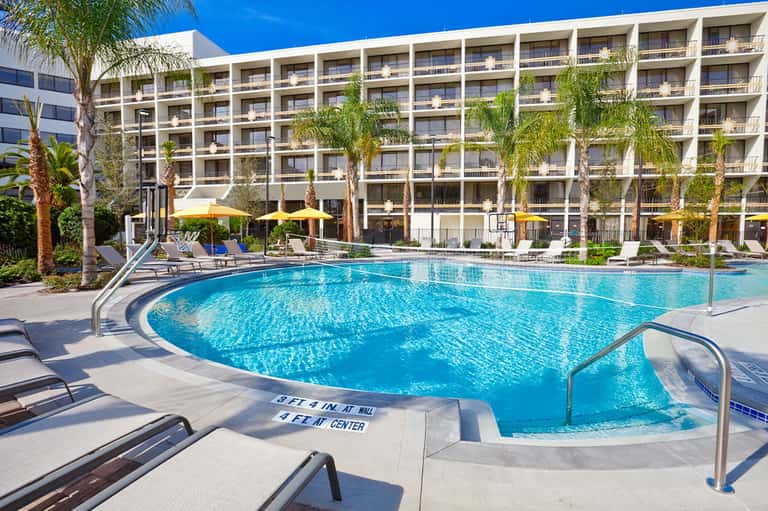 Large resort-style pool with palm trees, yellow umbrellas, lounge chairs, and multi-story hotel building