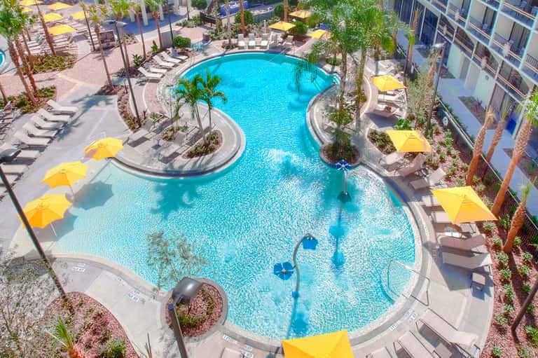 Aerial view of curved resort pool with yellow umbrellas, palm trees, and surrounding buildings